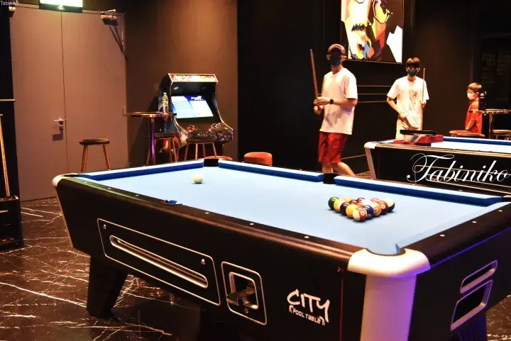 Modern pool table in a dimly lit entertainment lounge with arcade games and patrons playing billiards.