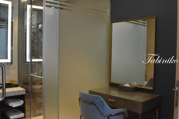 Modern hotel bathroom vanity with a gold-framed mirror, blue velvet chair, and glass shower enclosure.