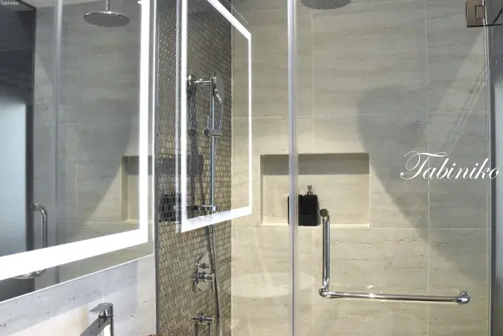 Modern bathroom with a glass shower enclosure, backlit mirror, and beige stone tiles.