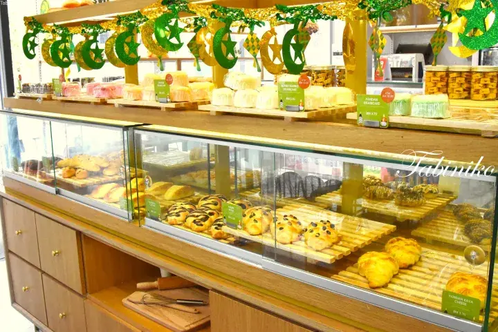 Bakery display case with bread and cakes, decorated with green and gold Hari Raya festive ornaments.