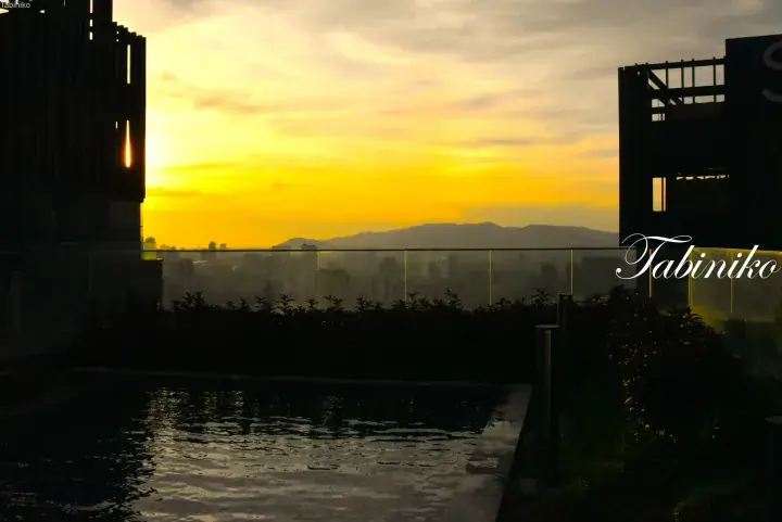 Sunset view from a rooftop infinity pool overlooking a city skyline and distant mountains.