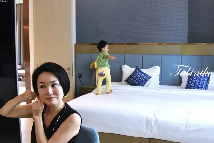 Woman adjusting earrings in a modern hotel room while a toddler plays on the bed.