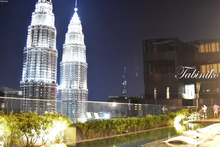 Night view of Petronas Twin Towers from a rooftop pool deck with modern lounge chairs and greenery.