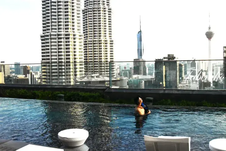 Man relaxing in a rooftop infinity pool overlooking the Kuala Lumpur skyline, featuring Petronas Towers and KL Tower.
