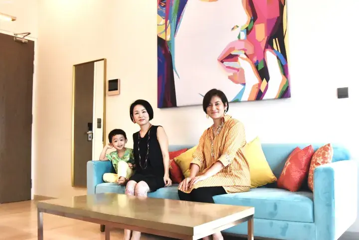 Two women and a young boy sitting on a blue sofa in a modern apartment living room with a large colorful painting on the wall.
