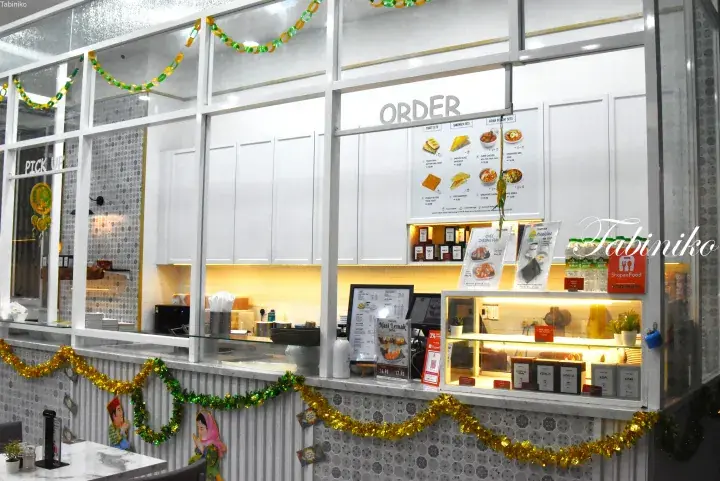 Modern white food stall counter with menu boards, display cases, and festive green and gold decorations.
