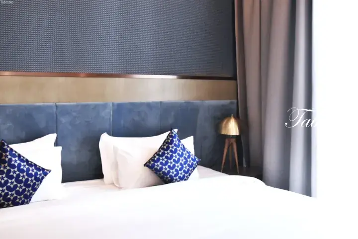 Modern hotel bedroom featuring a blue velvet headboard, white linens, and decorative blue patterned cushions.