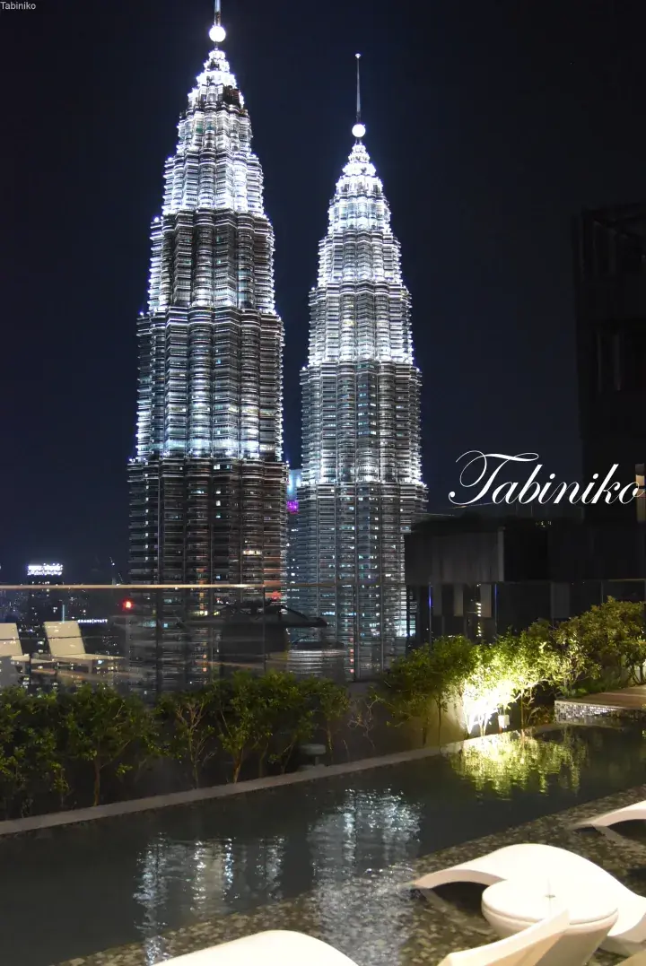 Petronas Twin Towers illuminated at night, viewed from a rooftop infinity pool with lounge chairs and greenery.