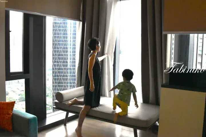 Woman and toddler looking out from a high-rise apartment window with a view of a modern building.