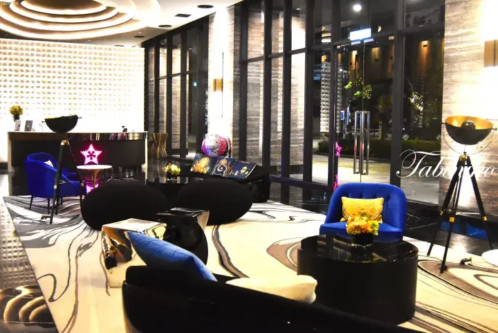 Modern hotel lobby featuring blue velvet chairs, black sofas, and a textured white wall with ambient lighting.