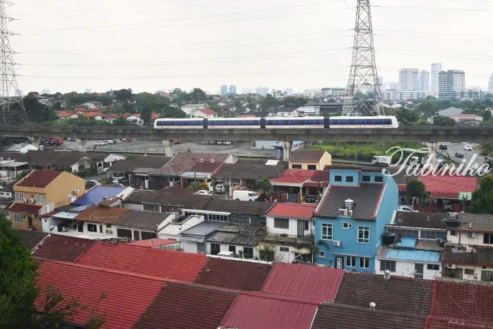 Rapid KL train on elevated track passing over residential rooftops with city skyline in background.