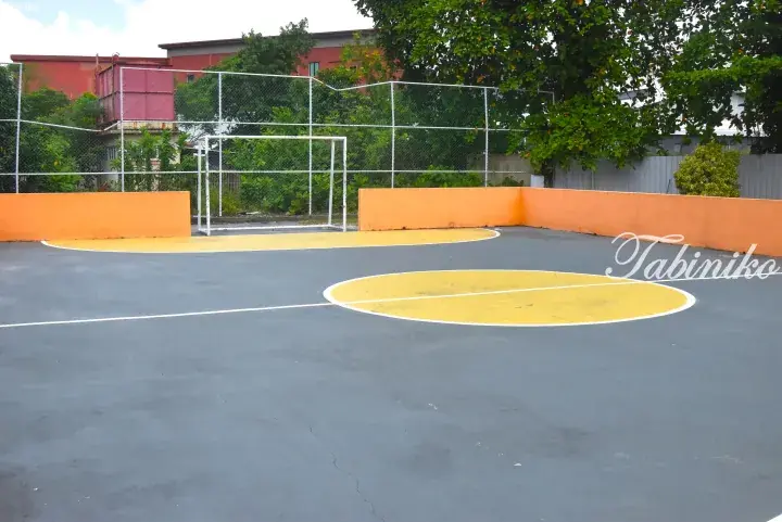 Outdoor sports court with grey asphalt, yellow markings, orange walls, and a white soccer goal.