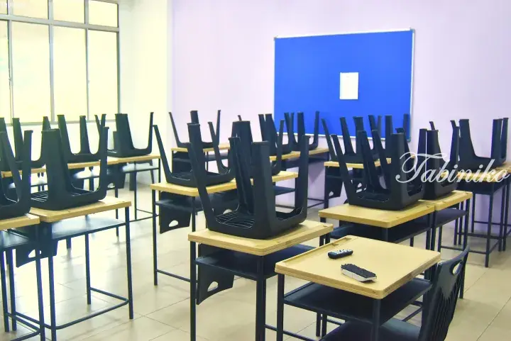 Empty classroom with black chairs stacked upside down on wooden desks, a blue bulletin board, and a remote on one desk.