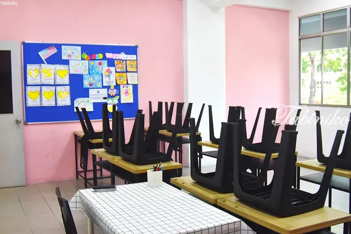 Empty classroom with chairs stacked on desks, pink walls, and a blue bulletin board displaying children’s artwork.