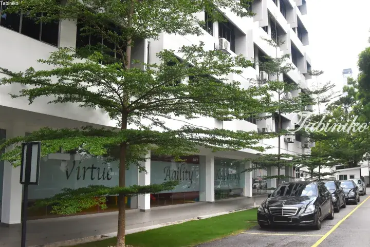 Modern white building exterior with glass storefronts displaying ‘Virtue’, ‘Agility’, and ‘Knowledge’ words, shaded by green trees.