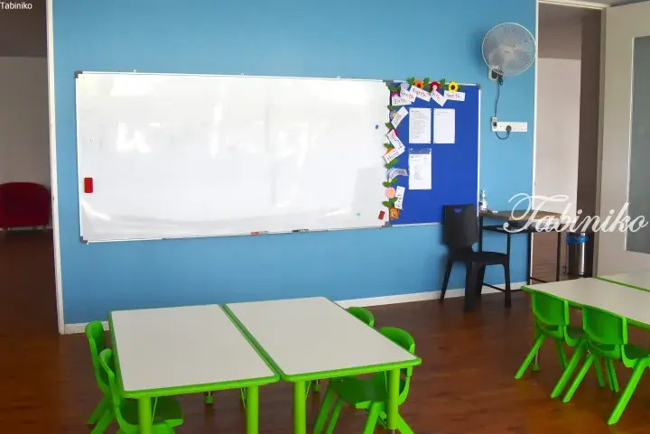 Bright classroom with green children’s tables and chairs, whiteboard, and blue wall.