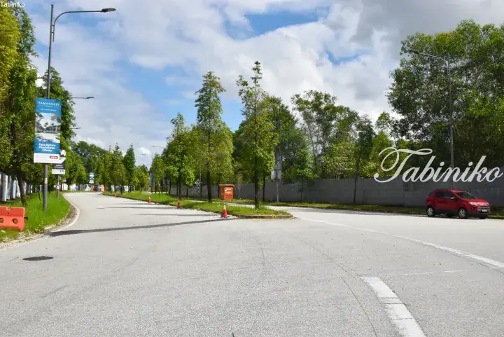 A quiet road in the new Tamansari Sarang development with trees, a red car, and a banner announcing new registration.