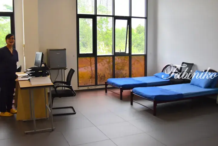 Staff member in a bright clinic room with two blue examination beds and large windows overlooking greenery.