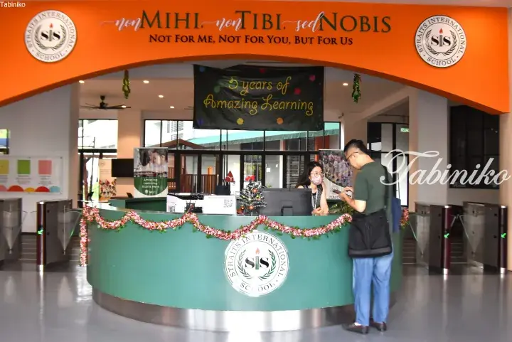 Straits International School reception desk with orange archway displaying motto ’non MIHI, non TIBI, sed NOBIS’ and a banner celebrating 5 years of learning.
