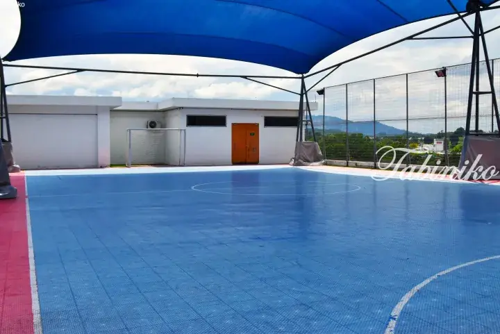 Blue modular sports court with red borders under a shade canopy, featuring a soccer goal and white building.