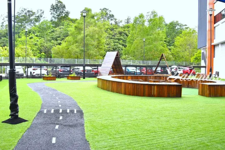 Modern playground with curved wooden seating and a winding path on green turf, set against a backdrop of trees and a parking lot.