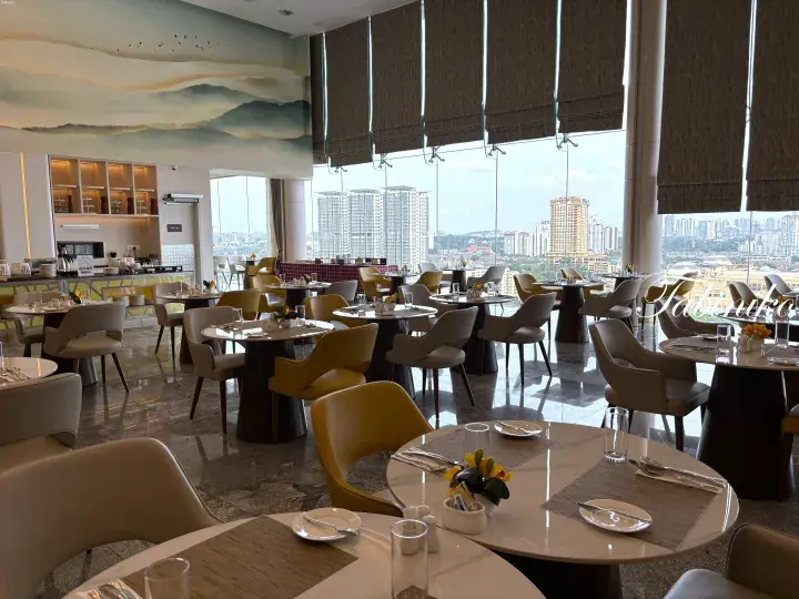 Modern hotel dining area with round tables, yellow chairs, and panoramic city skyline views through floor-to-ceiling windows.