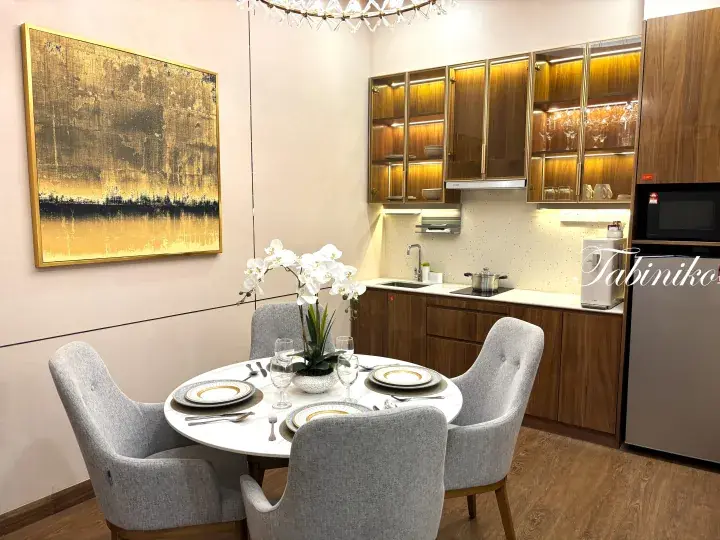 Modern dining area with round table, grey chairs, and kitchenette featuring wooden cabinets and gold accents