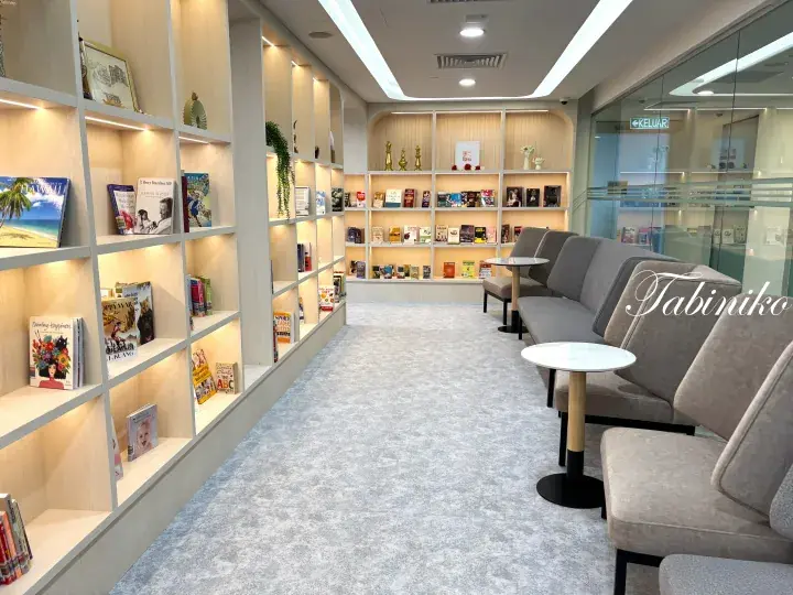 Modern library reading area with built-in wooden shelves, grey sofas, and small tables in a bright, carpeted space.