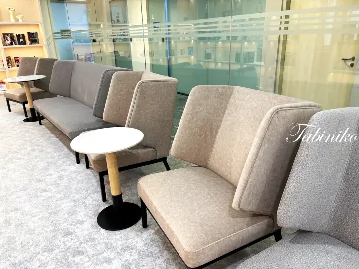 Modern beige armchairs and a sofa arranged in a bright lounge with small round tables and glass walls.