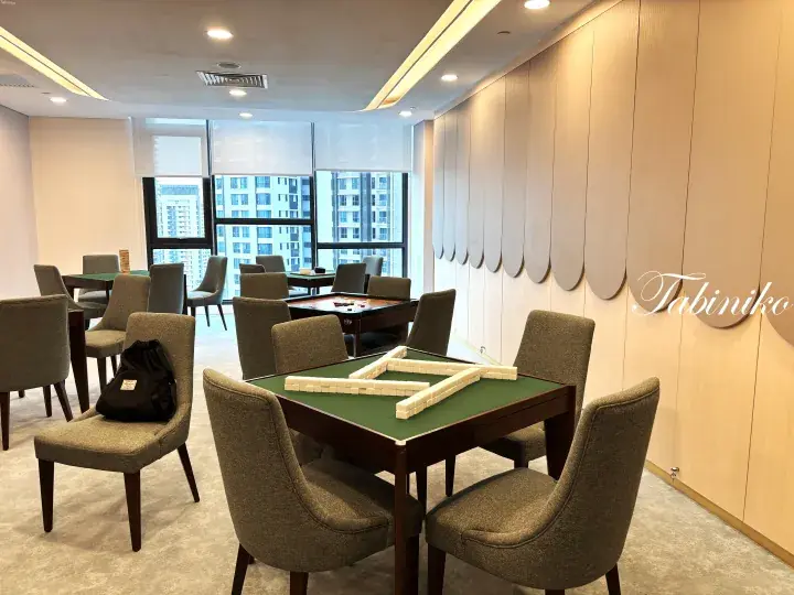 Modern mahjong room with wooden tables, green felt, and comfortable chairs near large windows.