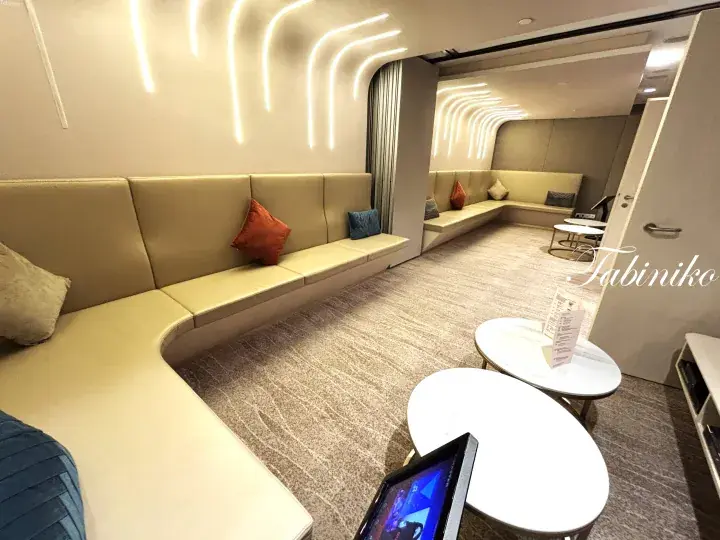 Modern airport lounge with curved beige seating, decorative LED wall lights, and small round tables.