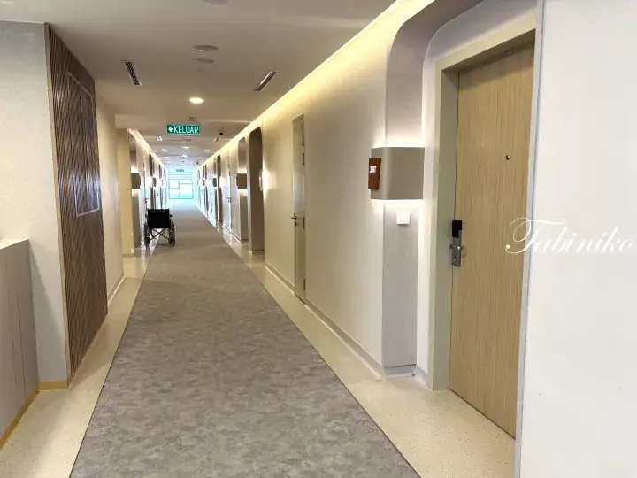 A modern, well-lit corridor in a healthcare facility with wooden doors and a wheelchair parked along the carpeted hallway.