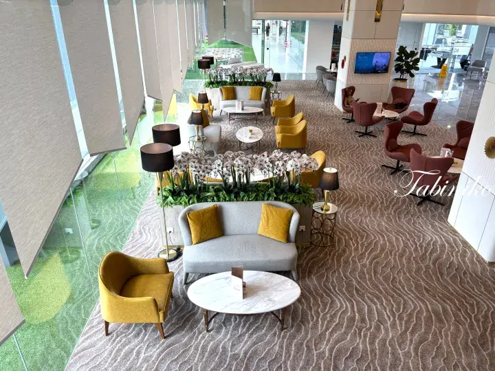 Modern hotel lobby with yellow and grey seating, marble tables, and large windows overlooking a green landscape.