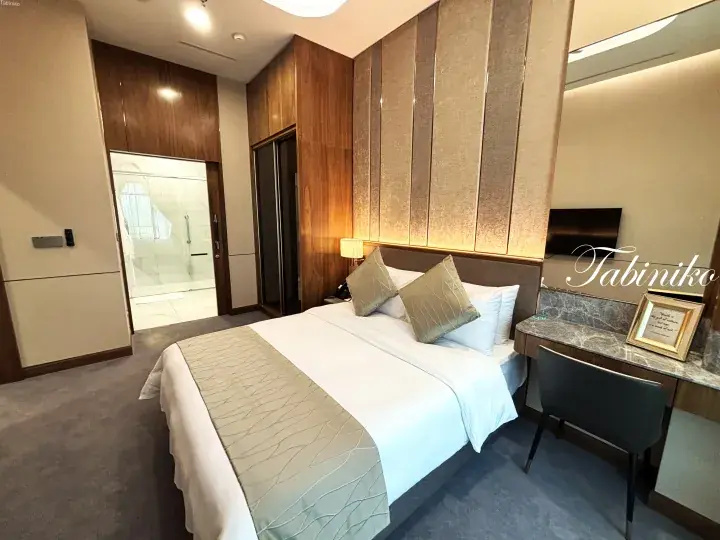 Modern hotel room with a neatly made bed, wooden accents, and a glass-enclosed bathroom visible through an open door.