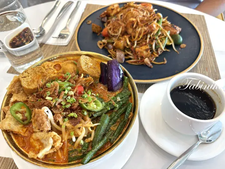 A bowl of laksa with tofu, egg, and vegetables alongside a plate of stir-fried noodles and kopi in a Malaysian restaurant.