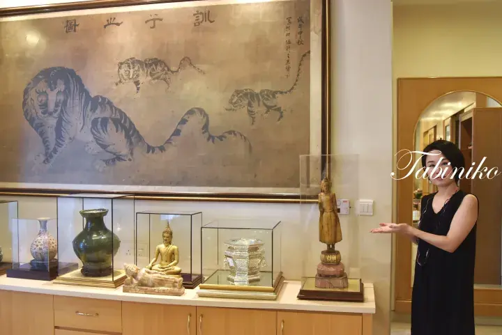 Woman presenting a display of antique vases and Buddha statues beneath a large traditional tiger painting in a Malaysian home.