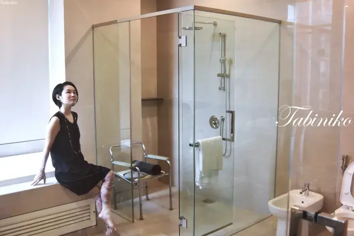Woman in black dress sitting on a bathtub ledge in a modern hotel bathroom with a glass shower enclosure.