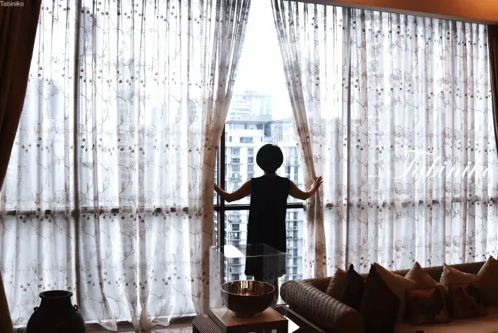 Woman opening sheer curtains to reveal a city view from a modern apartment balcony.