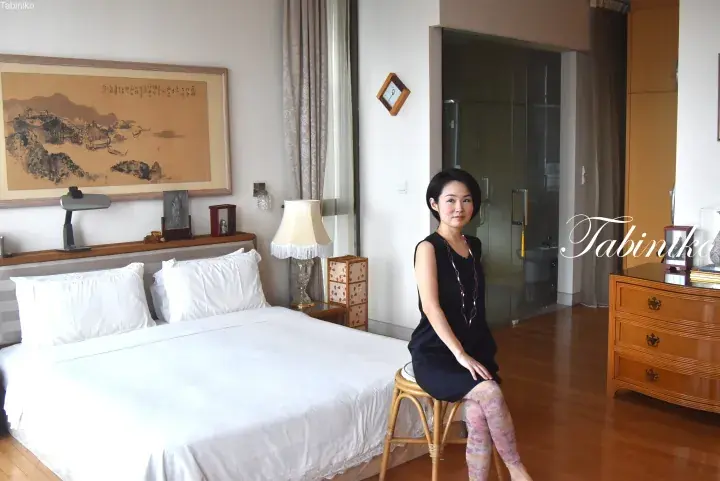 Woman sitting on a rattan stool in a hotel room featuring a large bed, traditional Chinese landscape painting, and glass-enclosed bathroom.