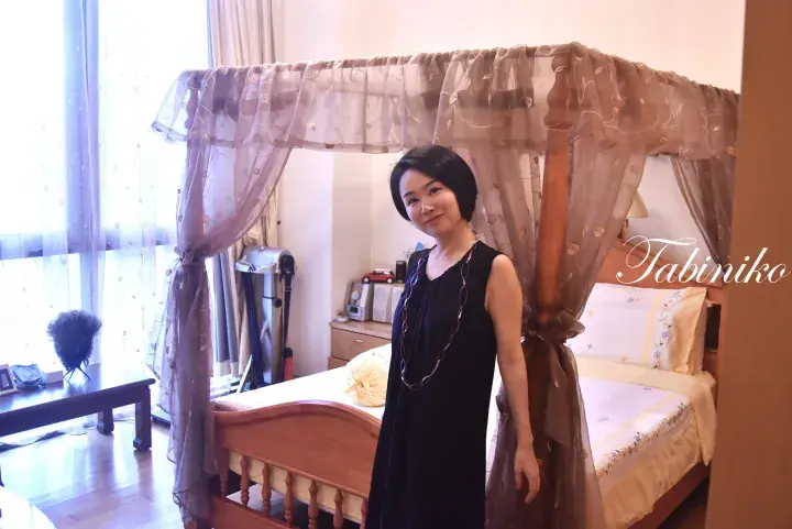 Woman in a black dress standing beside a wooden four-poster bed with sheer curtains in a bright bedroom.