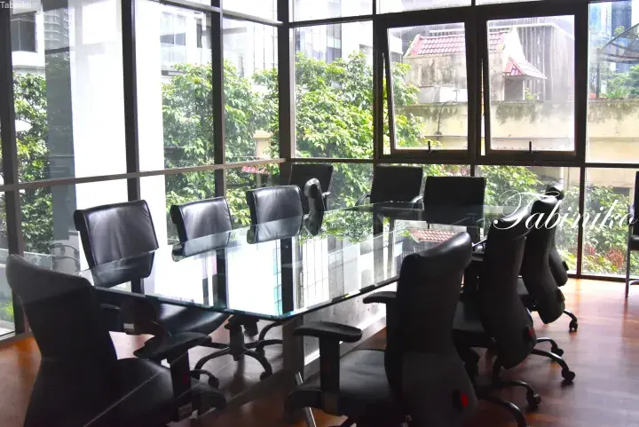 Modern office meeting room with a glass table and black chairs, featuring large windows with a view of lush greenery and nearby buildings.