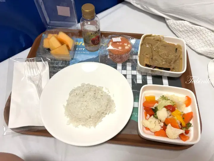 Hospitality tray with rice, mixed vegetables, curry, fruit, muffin, and drink on a bed.