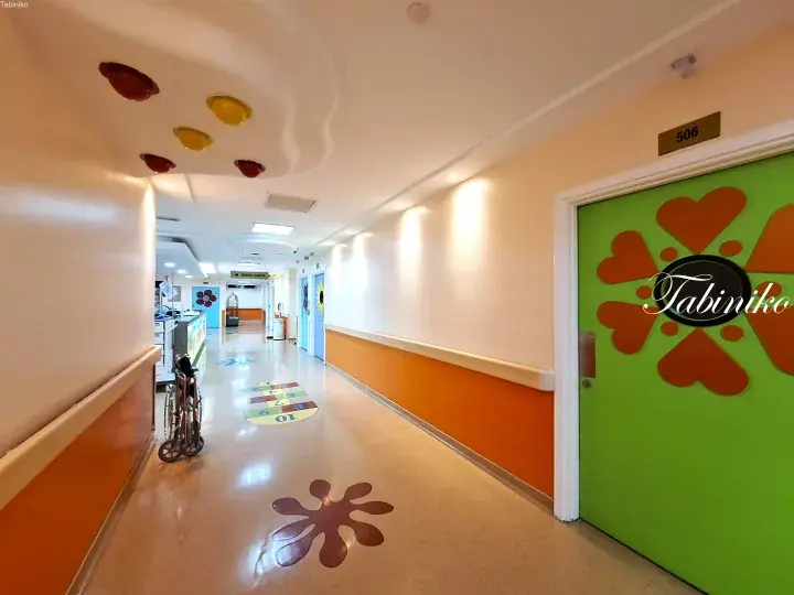 Colorful hospital corridor with playful hopscotch floor decals, heart-shaped door art, and a wheelchair parked along the wall.