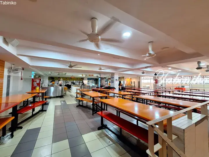 Spacious university cafeteria with orange tables and red benches, ceiling fans, and food stalls in the background.