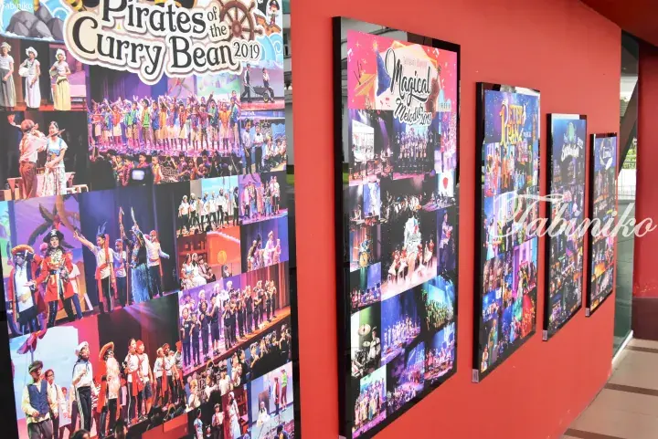 Red wall displaying large posters of children’s musical performances, including Pirates of the Curry Bean 2019 and Magical Melodies.