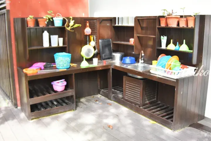 Outdoor wooden play kitchen with colorful plastic dishes, sink, and shelves for children’s imaginative play.