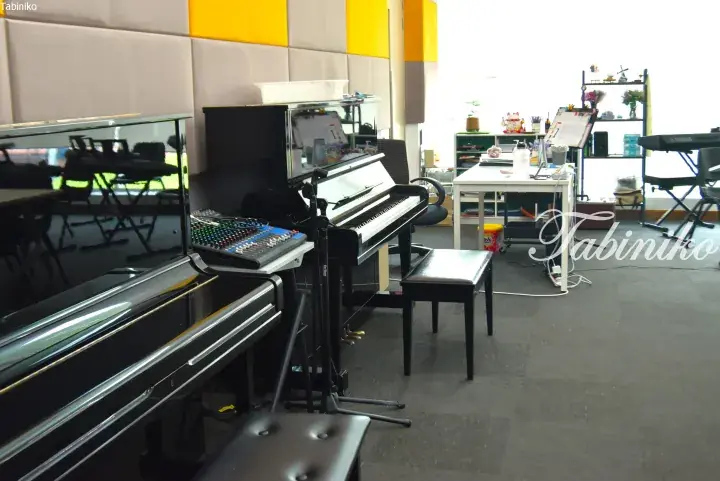 Interior of a music studio featuring a black grand piano, upright piano, and mixing console in a bright room.