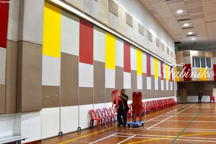 Staff moving red chairs in a large hall with colorful acoustic wall panels and a wooden sports court floor.