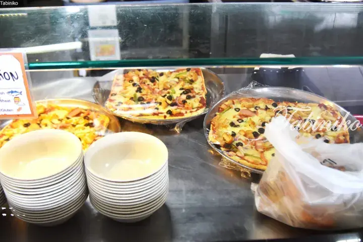 Assorted Malaysian pizzas displayed in a glass case with stacks of white bowls and a plastic bag nearby.
