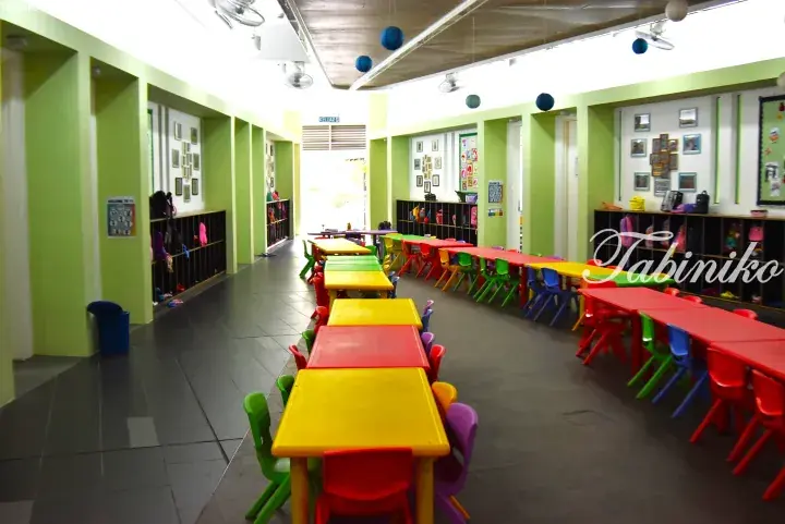 Bright classroom with colorful tables and chairs, green walls, and cubbies for a daycare or kindergarten setting.